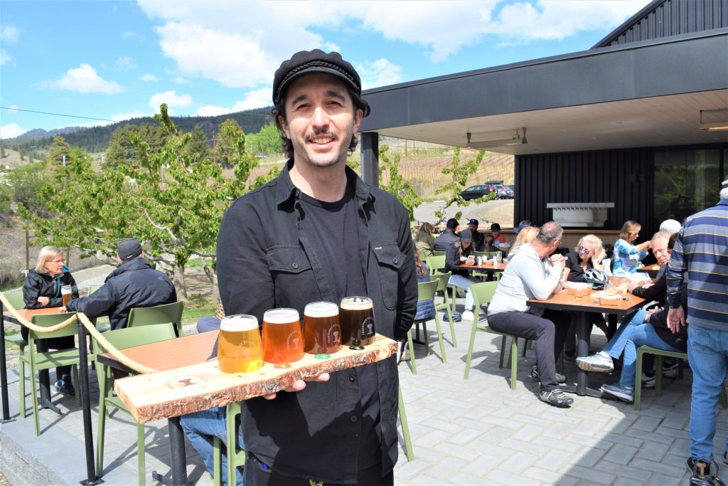 Josh from Abandoned Rail Brewing stands on the patio of the brewery wearing a black hat and shirt. He is holding a beer flight with 4 different beers arranged from lightest to darkest.
The patio tables are full with customers and it is sunny.