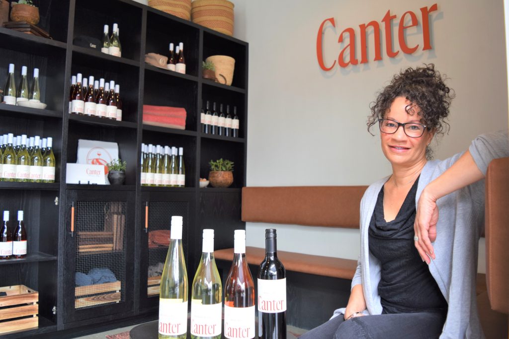 A woman is seen sitting next to four bottles of wine - 2 white, 1 rose, 1 red.
In the background the text 'Canter' is seen on the wall next to a cabinet featuring the wines for sale.