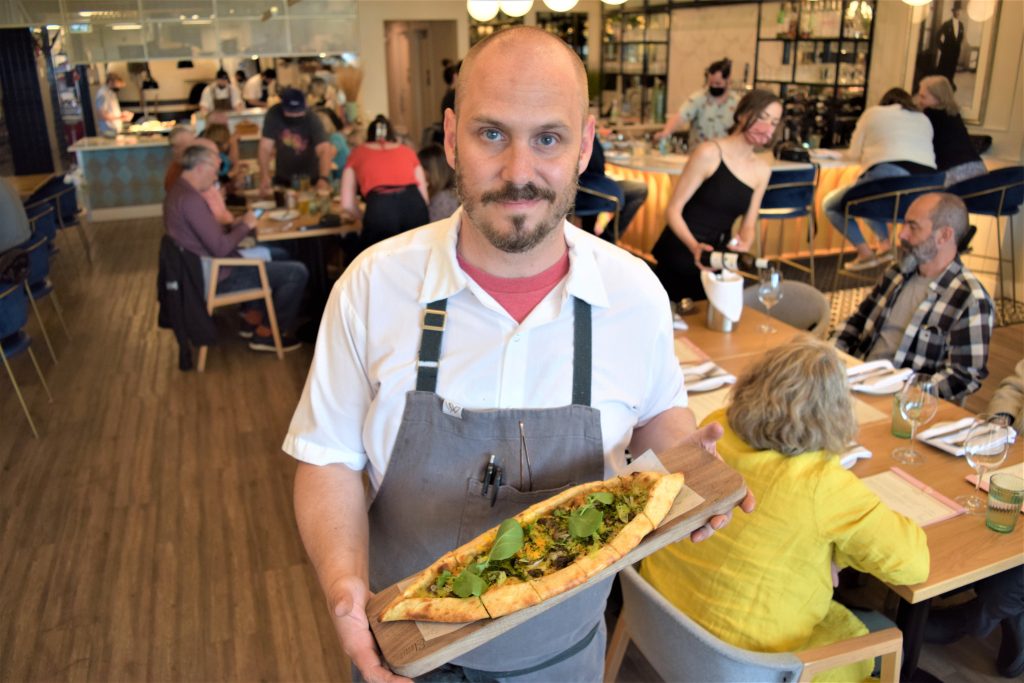 A chef at Elma restaurant is pictured holding a boat-shaped flatbread. 

He is wearing a white shirt with an apron and is standing in the restaurant. 

The restaurant tables are full and wine is being served. The busy kitchen and bar are also in view. 
