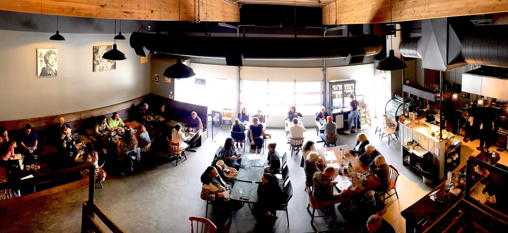 The restaurant Lokis Garage is pictured from a raised perspective. 

There are customers at every table eating and the garage doors in the background are bright with sunlight. 