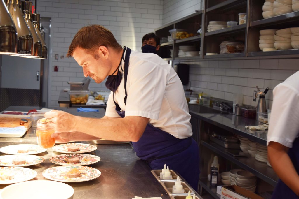 Chef Ned Bell from the Naramata Inn is seen in the kitchen standing over 7 plates on the stainless steel counter.
He is adding a garnish to the dishes.