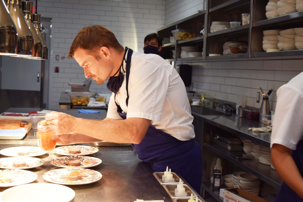 Chef Ned Bell from the Naramata Inn is seen in the kitchen standing over 7 plates on the stainless steel counter. 
He is adding a garnish to the dishes. 