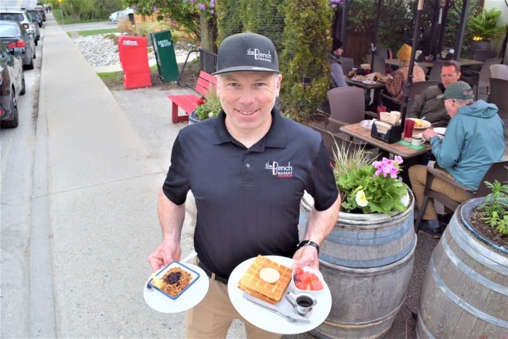 A man is pictured in front of the patio at The Bench Market. He is wearing a branded hat and shirt and holding one plate in each hand.
In his right hand is a dish of granola with fruit on top and in his left hand is a waffle with syrup and fruit on the side.
In the background, the patio tables in view have customers eating at them.