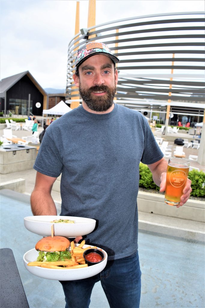 A man holding a beer in one hand and a burger dish and pasta bowl in the other.
He has a hat and a beard and is wearing a t shirt and jeans.
He is standing at the District Wine Village.
