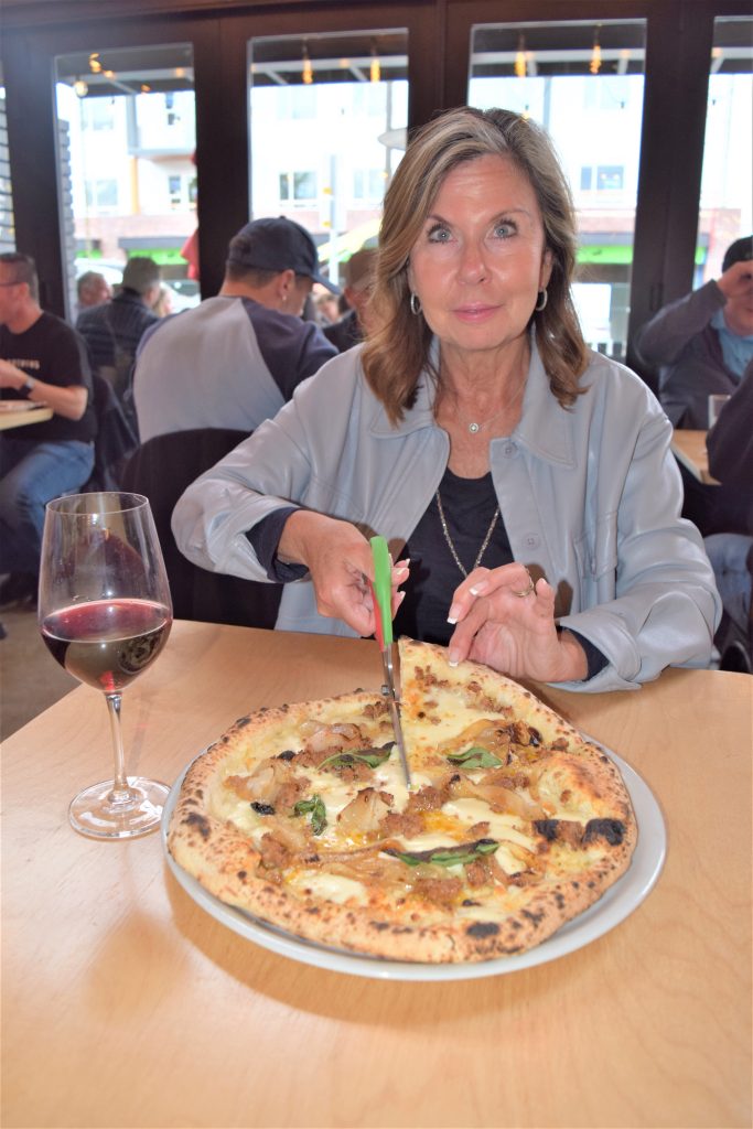 A woman is holding a pair of scissors as she cuts her pizza.
She has a glass of red wine on the table next to her.
In the background the other tables are seated with customers in front of the large glass windows.