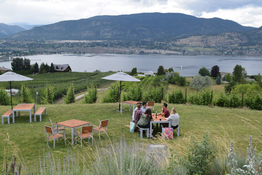 A grassy patio area at Poplar Grove Winery is pictured with 4 tables - one table has 5 people sitting at it. 

The grassy area overlooks vineyards and Okanagan Lake beyond the grape vines. 