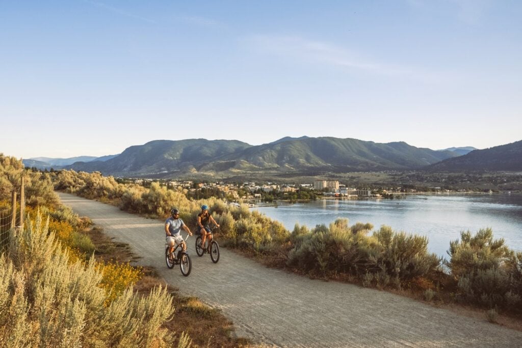 A man and a woman are pictured on ebikes on the KVR trail above lake okanagan.