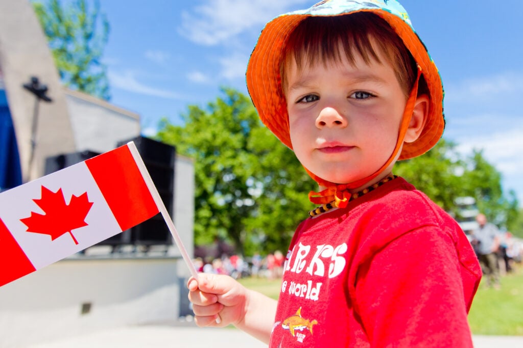  A young boy holding a Canada flag is pictured wearing a bucket hat and red t shirt.