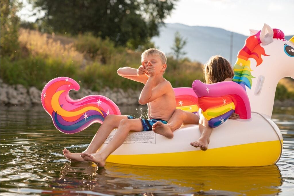 Two children on a unicorn floatie are pictured. the boy is pointing finger guns at the camera and the girl is not looking.