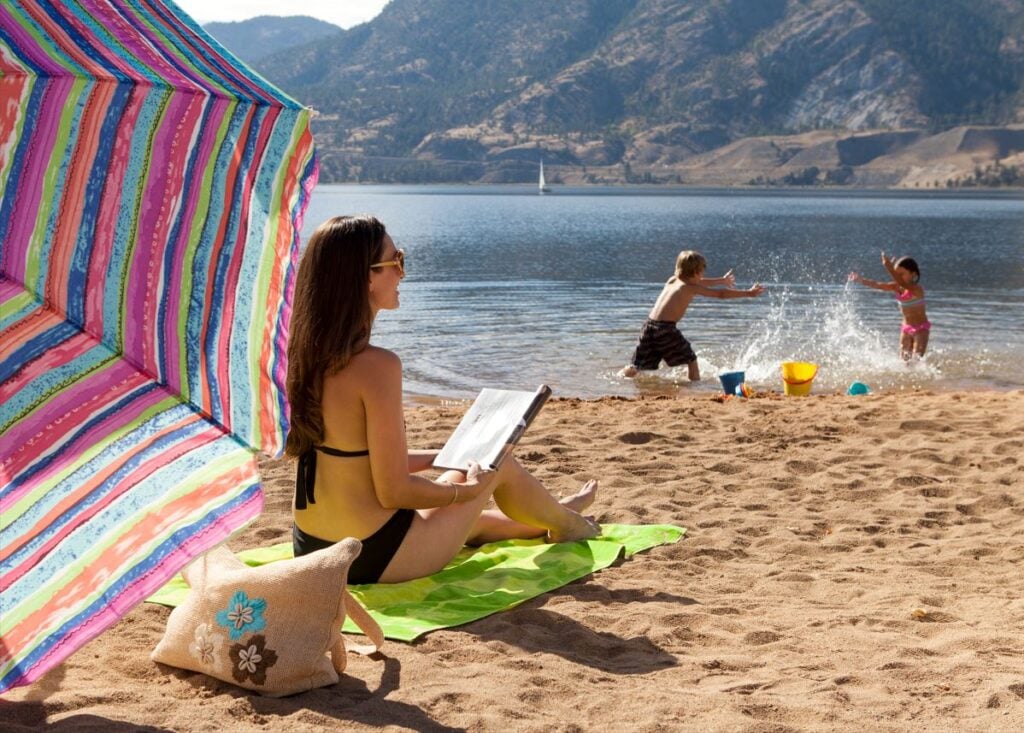 A woman sitting on the beach under an umbrella whie her two kids splash in the lake